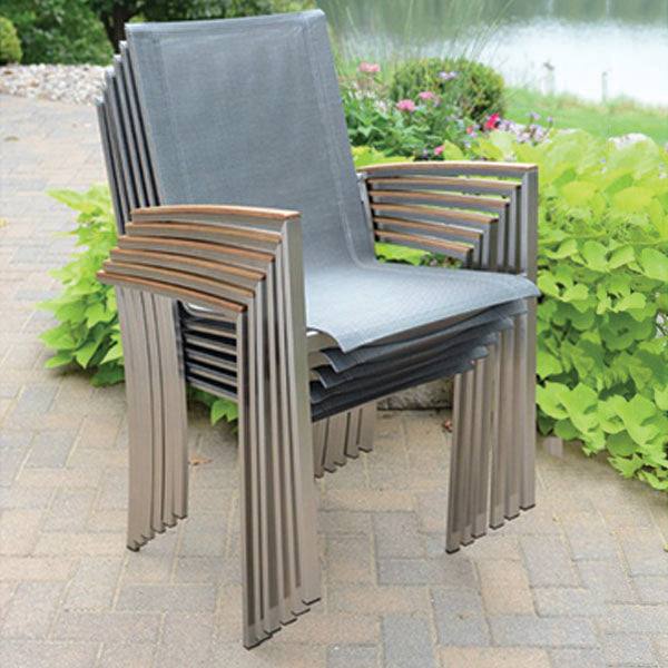 The Avanti Outdoor Patio Teak Dining Set by Delivery Only features four stackable chairs with ergonomic gray mesh sling seats, wooden armrests, and stainless steel frames. This set graces a stone patio against a backdrop of greenery, flowers, and a tranquil pond.