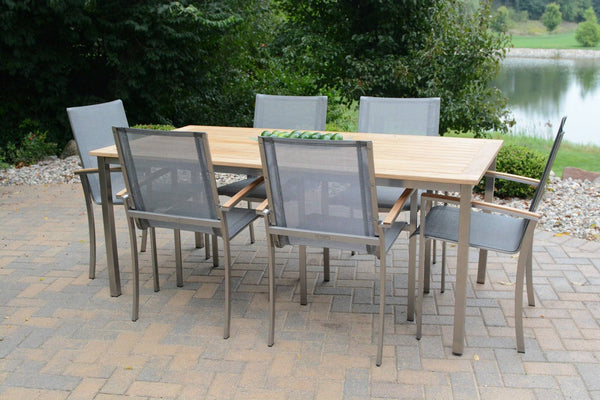 An Avanti Outdoor Patio Teak Dining Set by Delivery Only graces the stone patio, featuring a wooden table and six gray mesh chairs with ergonomic sling seats. The serene setting, complemented by the lush trees and pond, ensures a perfect outdoor dining experience.