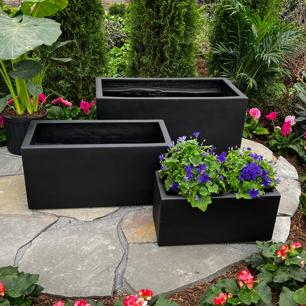 Three Pick-Up & Local Delivery Only Berkley Trough Planters (30x15, black) sit on a stone patio; one holds purple flowers and greenery, while the other two are empty. These UV-resistant planters are ideal for outdoor use.