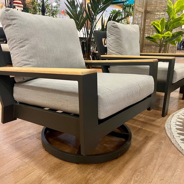 Two modern armchairs from the Delivery Only Bozania Outdoor Patio 4-Piece Set, with grey Sunbrella cushions and wooden armrests, sit on a wooden floor. They have a dark metal frame and circular base, surrounded by lush green plants and a decorative mat, exemplifying outdoor luxury.