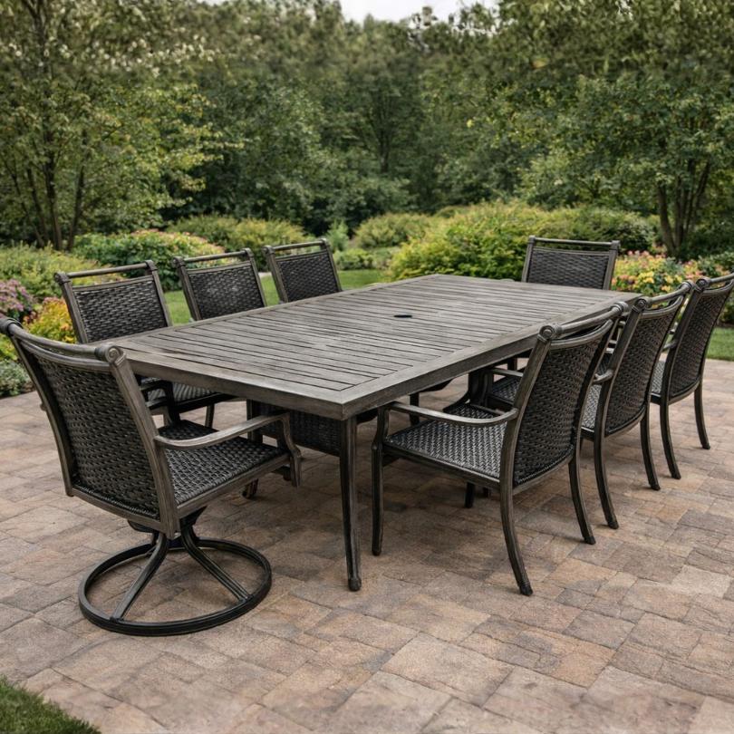 A Delivery Only Cabo 9-piece Outdoor Patio Dining Set, featuring eight black all-weather wicker chairs, is arranged on a paved patio with lush gardens and trees in the background.