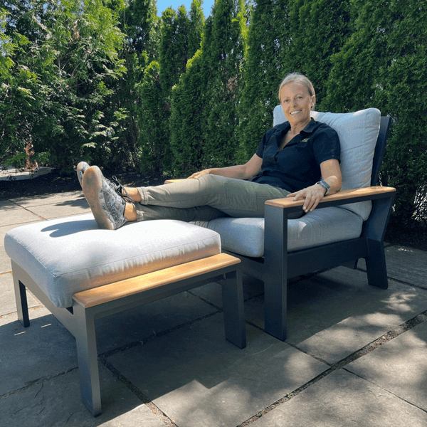 A person is enjoying a sunny day lounging on the Bozania Patio Lounge Chair Recliner by Delivery Only. Theyre smiling, dressed casually, with their legs up on an ottoman amid lush greenery and stone paving.