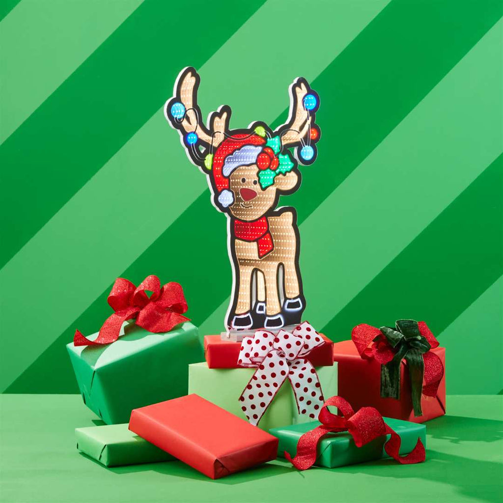 The Infinity Light – Reindeer - 24" from Available for Pick-Up & Delivery features a festive reindeer in a Santa hat and red scarf, standing among holiday gifts, perfect for adding seasonal charm to your home décor.
