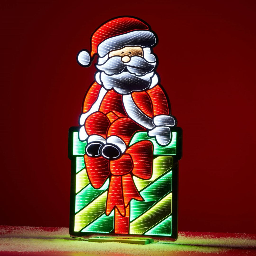 The Infinity Light – Santa with Package - 47" from Available for Pick-Up & Delivery features Santa sitting on a large, striped gift with a red bow, perfect for brightening your holiday décor.