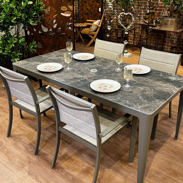 The Horizon Outdoor Patio Dining Set - 5 pc. from Delivery Only features a sintered stone table with a marble-patterned top and is elegantly set with five white plates and glasses, surrounded by five beige chairs in a chic indoor setting with brick walls and plants, featuring UV-resistant durability for outdoor use.