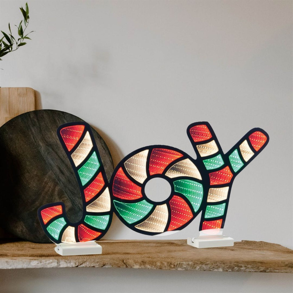 The Infinity Light – Joy – 24” by Available for Pick-Up & Delivery features red, green, and white lights, illuminating a wooden shelf and adding cheerful holiday decor next to a cutting board and a plant.
