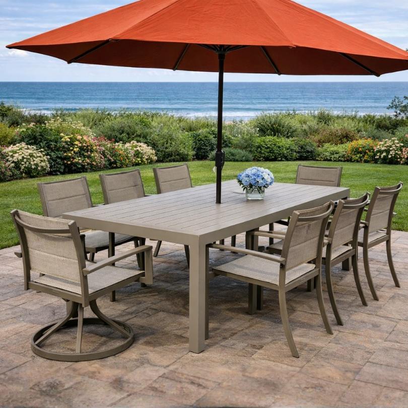 The Peak 9-piece Outdoor Patio Dining Set by Delivery Only features a durable aluminum frame, eight beige chairs, and a rectangular table with a blue flower arrangement and large orange umbrella—perfect for garden or ocean views.