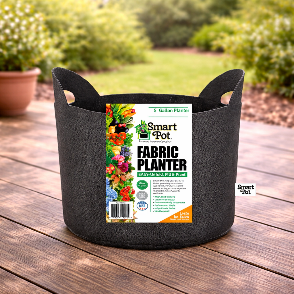 A black 5-gallon “Planter - Smart Pot with Handle” by Pickup & Local Delivery Only rests on a wooden deck, its label showing colorful flowers. In the background are blurred greenery and another root-pruning planter.