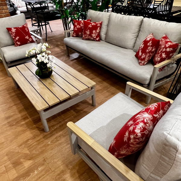 Enhance your outdoor space with the Titan 4-Piece Set by Delivery Only. This modern gray-cushioned patio sofa, lounge, and swivel chair set comes with red floral pillows. It includes a wooden coffee table adorned with a potted orchid, all amidst lush greenery on a polished floor.