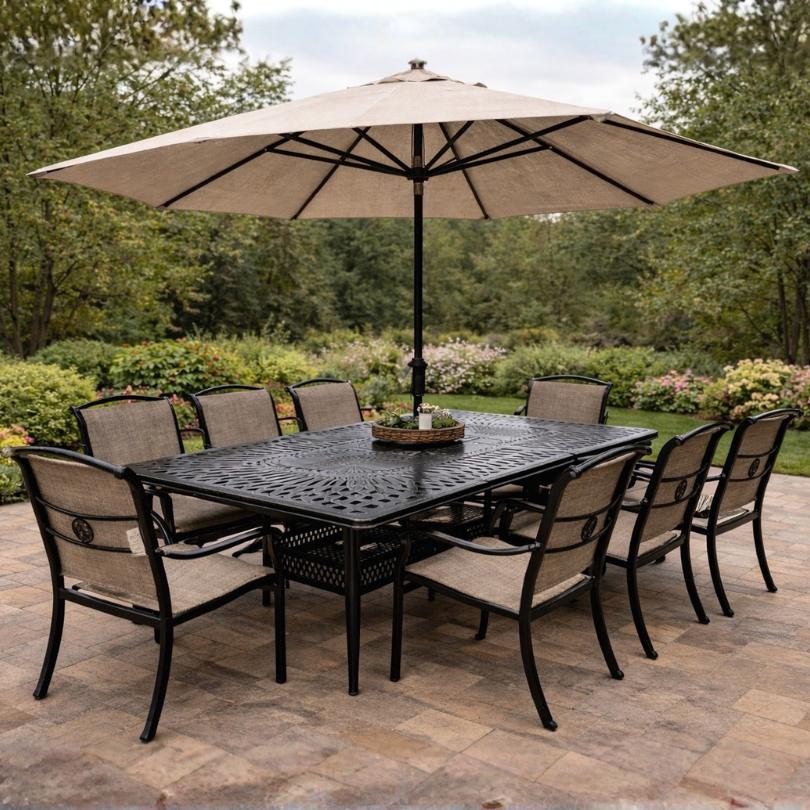 The Delivery Only Toscana 9-piece Outdoor Patio Extension Dining Set includes a black metal expandable table, eight cushioned chairs, and a beige umbrella—perfect for outdoor dining on a stone patio among greenery.
