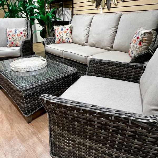 The Waverly Outdoor Patio Set by Delivery Only features a light gray all-weather wicker ensemble with premium cushions, including a sofa, lounge chair, swivel chair, and glass-top coffee table. Decorative floral pillows enhance the chairs amidst a serene backdrop of lush green plants.