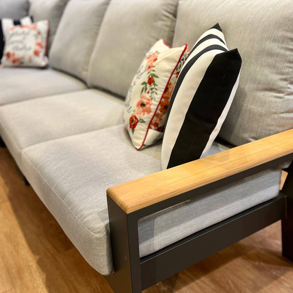 The Bozania Outdoor Patio Sectional Seating Set by Delivery Only features a modern gray design with light wooden armrests and decorative pillows in floral and striped patterns. Sunbrella® fabric cushions add style, while it sits perfectly on a wooden floor for a cozy ambiance.