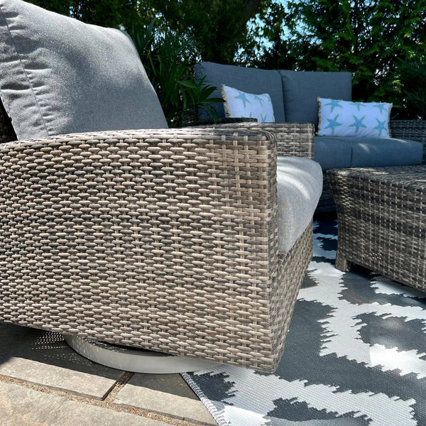 A close-up of Delivery Onlys Key Largo Outdoor Patio Set on a patio, featuring woven chairs with gray Sunbrella cushions. A matching loveseat with starfish-patterned pillows is in the background, set on a geometric-patterned rug amid leafy plants.