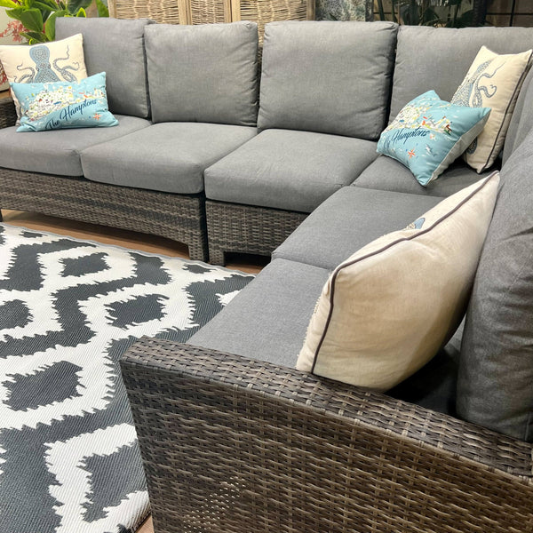 An inviting indoor seating space showcases the Key Largo Outdoor Patio Sectional from Delivery Only, with gray all-weather wicker and decorative blue and white Sunbrella® cushions. A patterned gray and white rug adorns the floor, complemented by charming woven baskets in the background.
