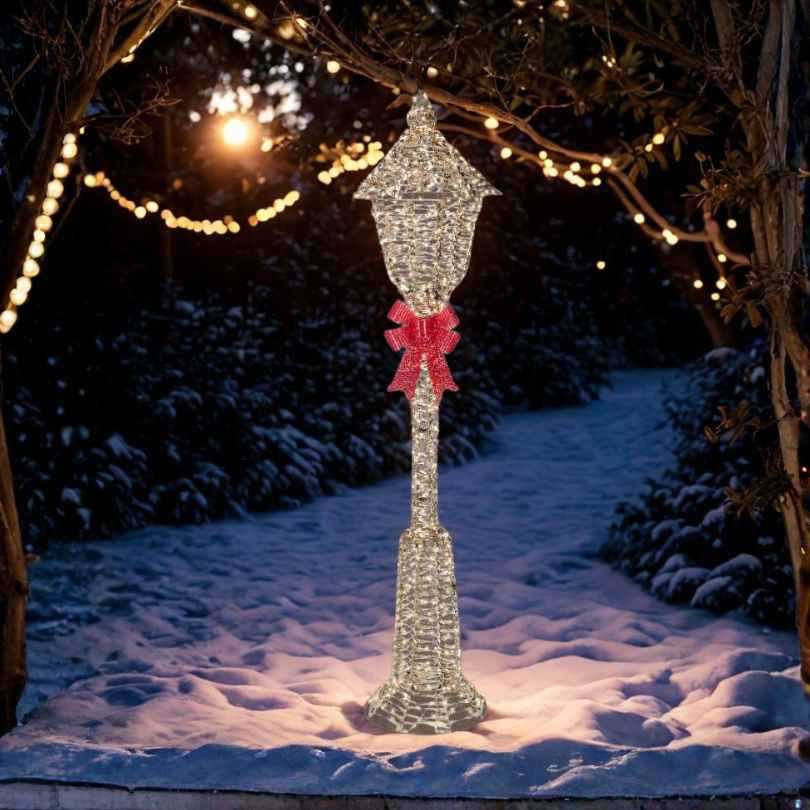 The Micro LED Lit Lamp Post with Red Bow – Warm White Lights by Available for Pick-Up & Delivery stands on snowy ground, creating the perfect outdoor Christmas decoration for your garden at dusk.