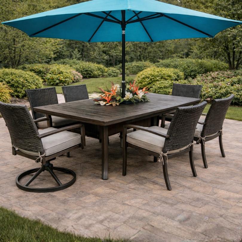 The Delivery Only Morena 7-piece Outdoor Patio Dining Set – B features six cushioned wicker chairs and a dark wood table. A large blue umbrella shades this stylish, weather-resistant set on a stone patio in a lush green garden.