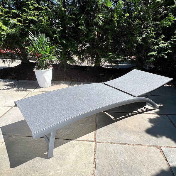 The Nettuno Outdoor Patio Chaise Lounge with Grey Sling by Pickup & Local Delivery Only is shown on a stone patio in sunlight, complemented by a potted plant and tall green shrubs in the background.