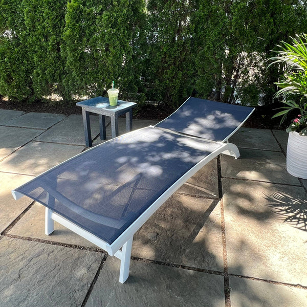The Nettuno Outdoor Patio Chaise Lounge with Navy Sling by Pickup & Local Delivery Only sits on a stone patio, its adjustable backrest facing sunlight and greenery, next to a side table with a green drink and potted plant nearby.