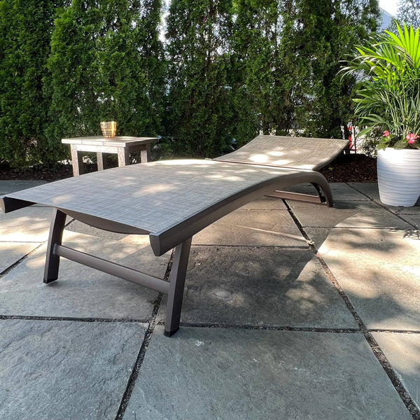 The Nettuno Outdoor Patio Chaise Lounge with Taupe Sling by Pickup & Local Delivery Only sits on a stone patio near green shrubs, a potted plant, and a small side table with a gold cup, while sunlight casts soft shadows.
