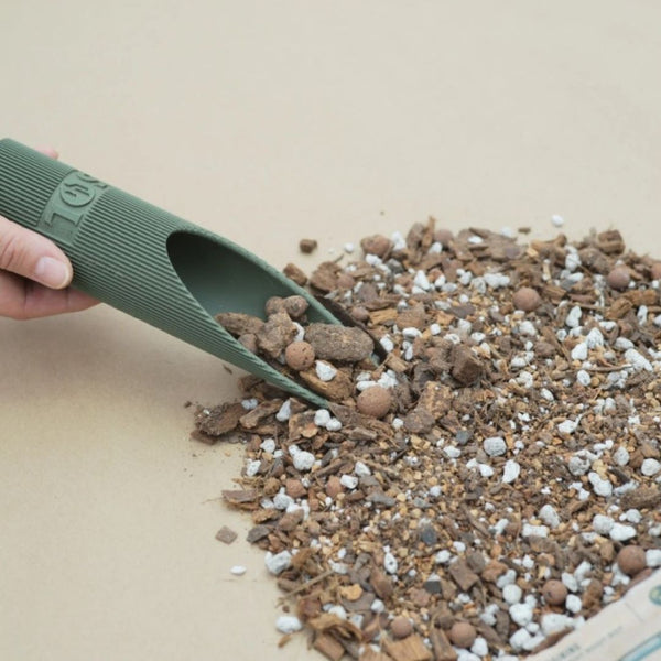 A hand uses a green scoop to pick up Sol Soil – Houseplant Chunky Mix (1 Gal.) by Available for Pick-Up & Delivery, featuring soil, pebbles, and perlite on a beige surface.