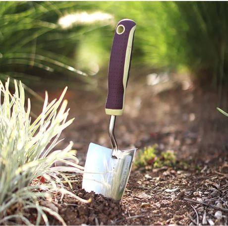 The Wilson & Ross - Trowel by All Delivery Options, featuring a purple and beige handle, is embedded in soil among green plants as sunlight highlights the stainless steel tool.