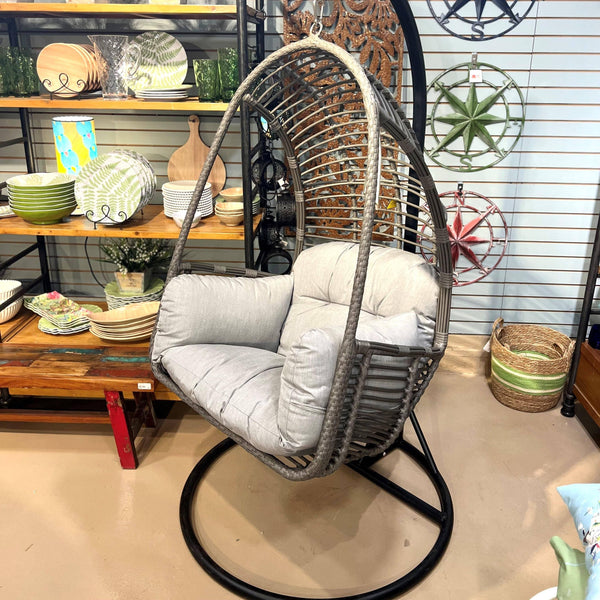 The Pick-Up & Local Delivery Only Hanging Basket Chair with a grey cushion is shown indoors near shelves filled with dishes, bowls, and decor, with wall art and baskets also visible in the room.