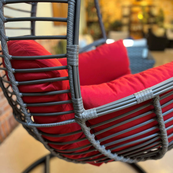 The Hanging Basket Chair by Pickup & Local Delivery Only features a durable woven rattan frame and vibrant red cushion, creating a cozy indoor seating spot with warm lighting and blurred furniture in the background.